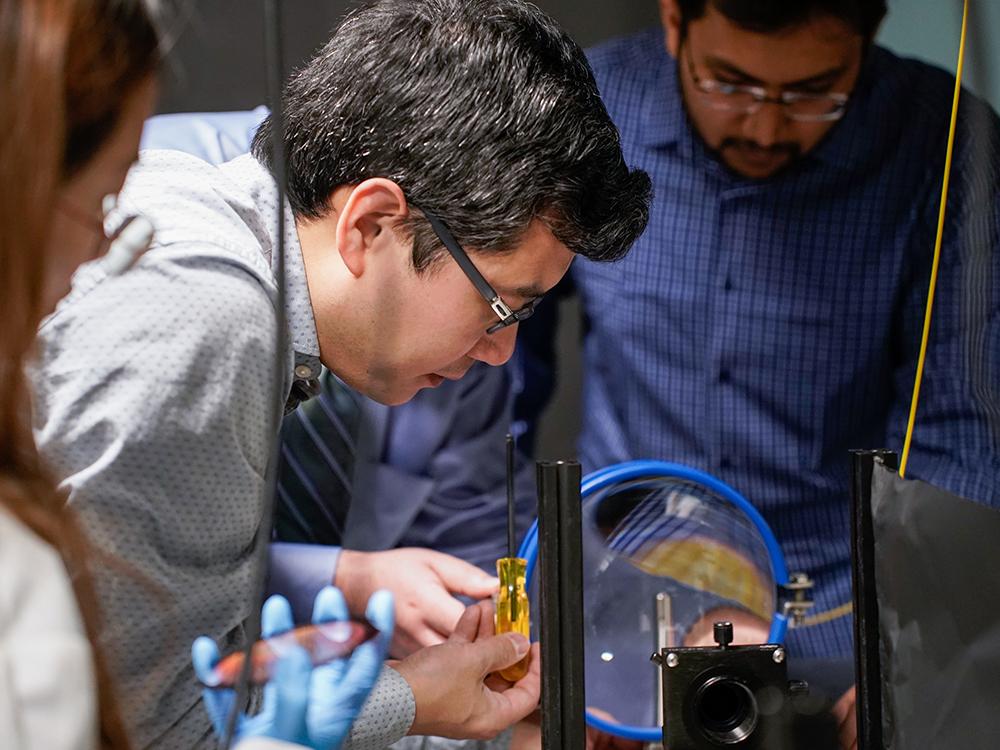 Xingjie Ni (center), Penn State associate professor of electrical engineering, manipulates a conventional telescope lens. In the foreground, Lidan Zhang, electrical engineering graduate student, holds the metalens prototype, which is much smaller, flatter, and more compact. Credit: Jeff Xu/Penn State .