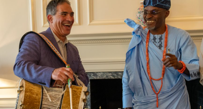 one man with an African drum laughing and standing next to a man in Nigerian garb
